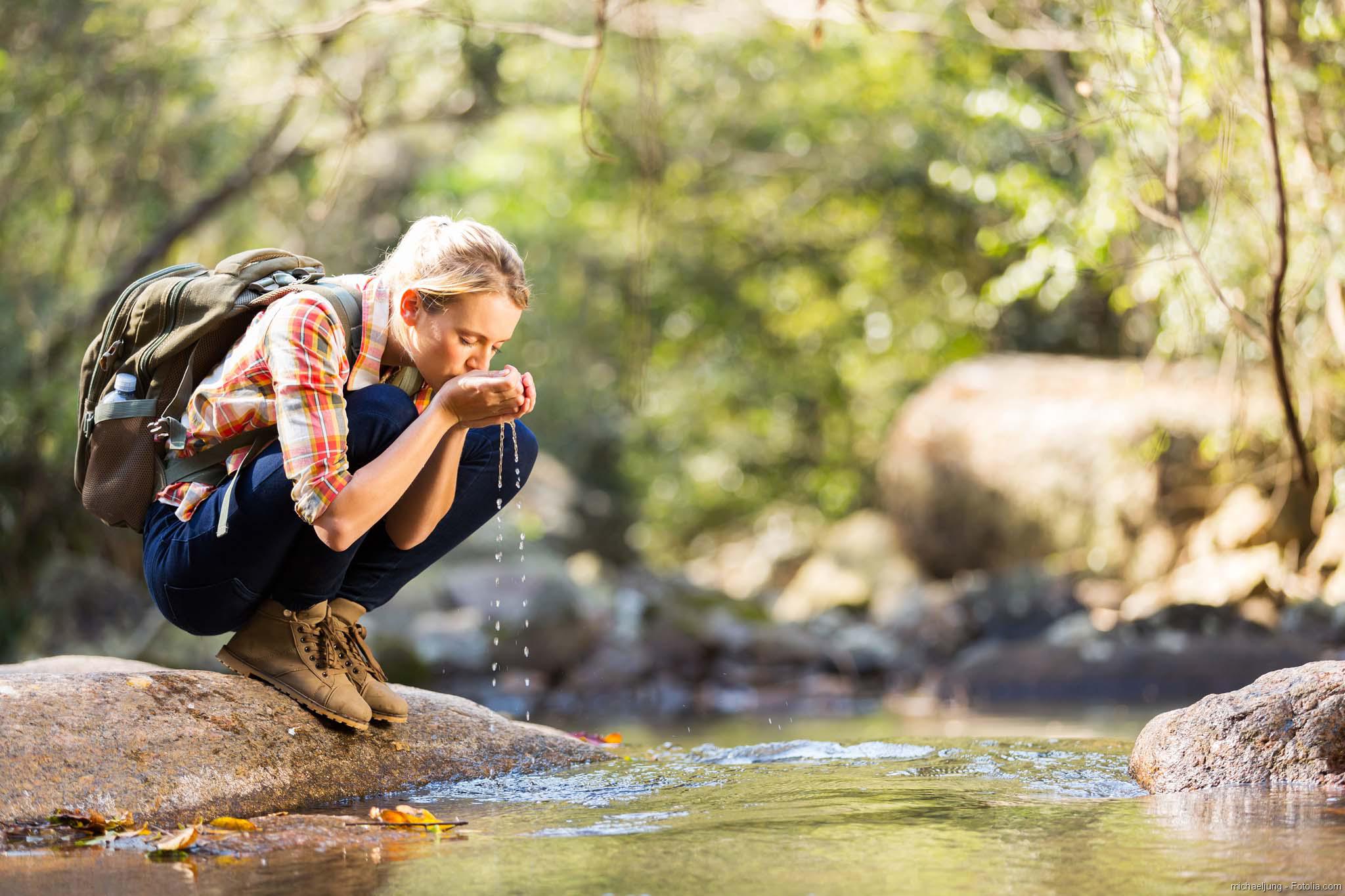 Junge Wanderin sitzt in der Hocke am Bach in den Bergen und trinkt Wasser